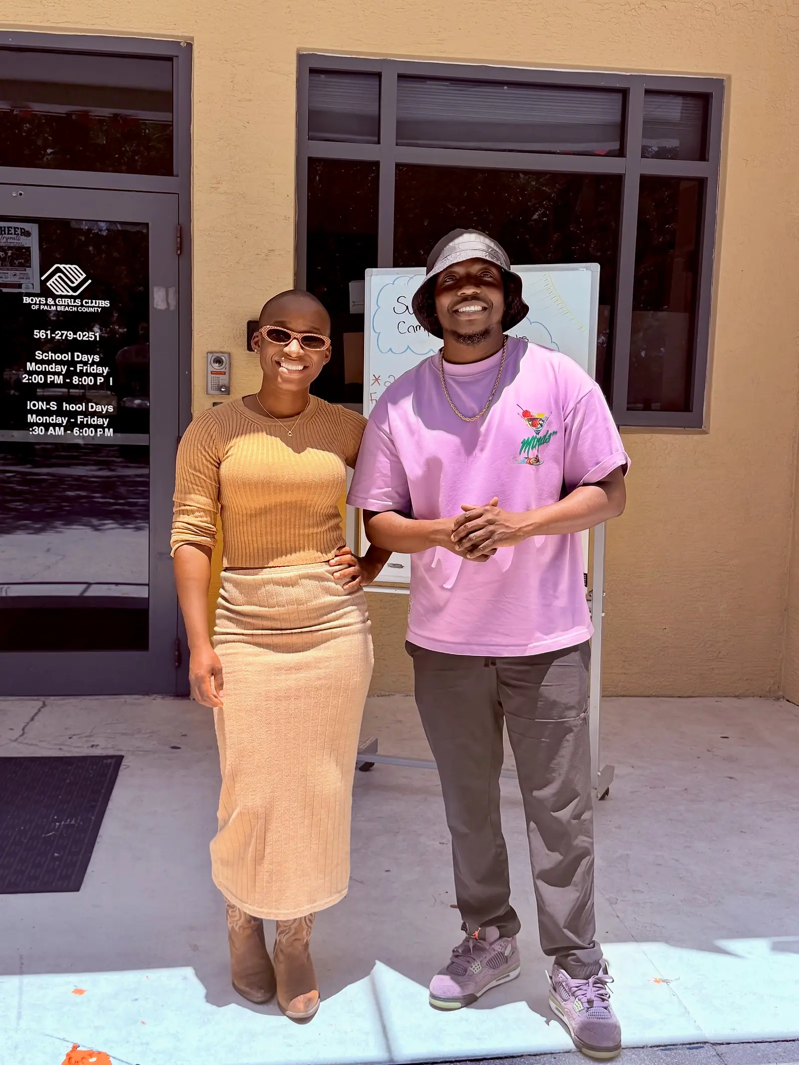 Two people standing outside a Boys & Girls Club building; the sign beside the door lists school-day and non-school-day opening hours.