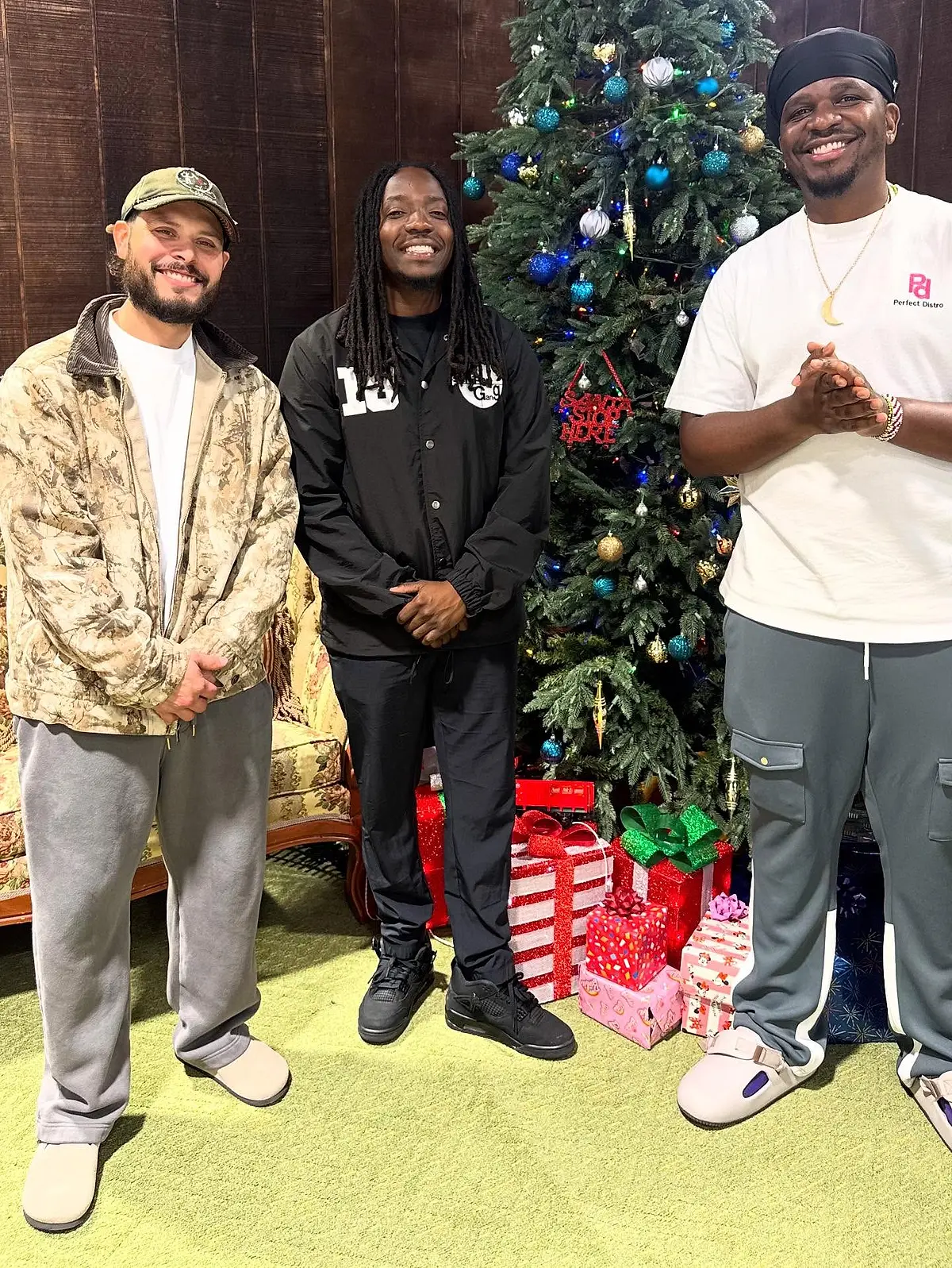 Three people standing beside a decorated Christmas tree with wrapped gifts at their feet.