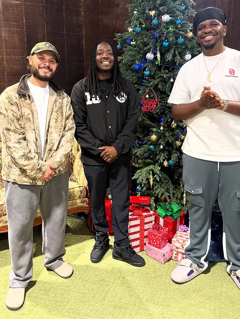 Three men standing in a room beside a decorated Christmas tree, wrapped gifts stacked on the floor in front of them.