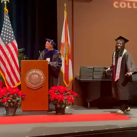 A speaker at a lectern between the United States and Florida flags, with a graduate in cap and gown nearby.
