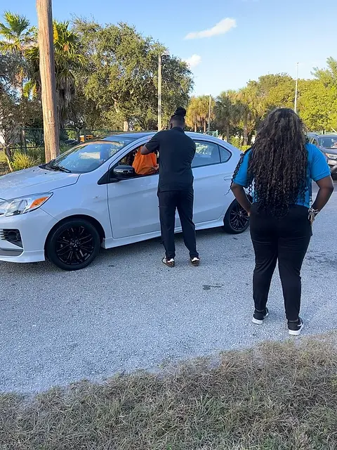 A car parked on a residential street with two people standing beside it in the shade.
