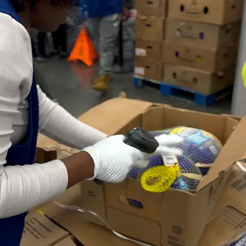 Gloved hands packing food into a cardboard box on a table.